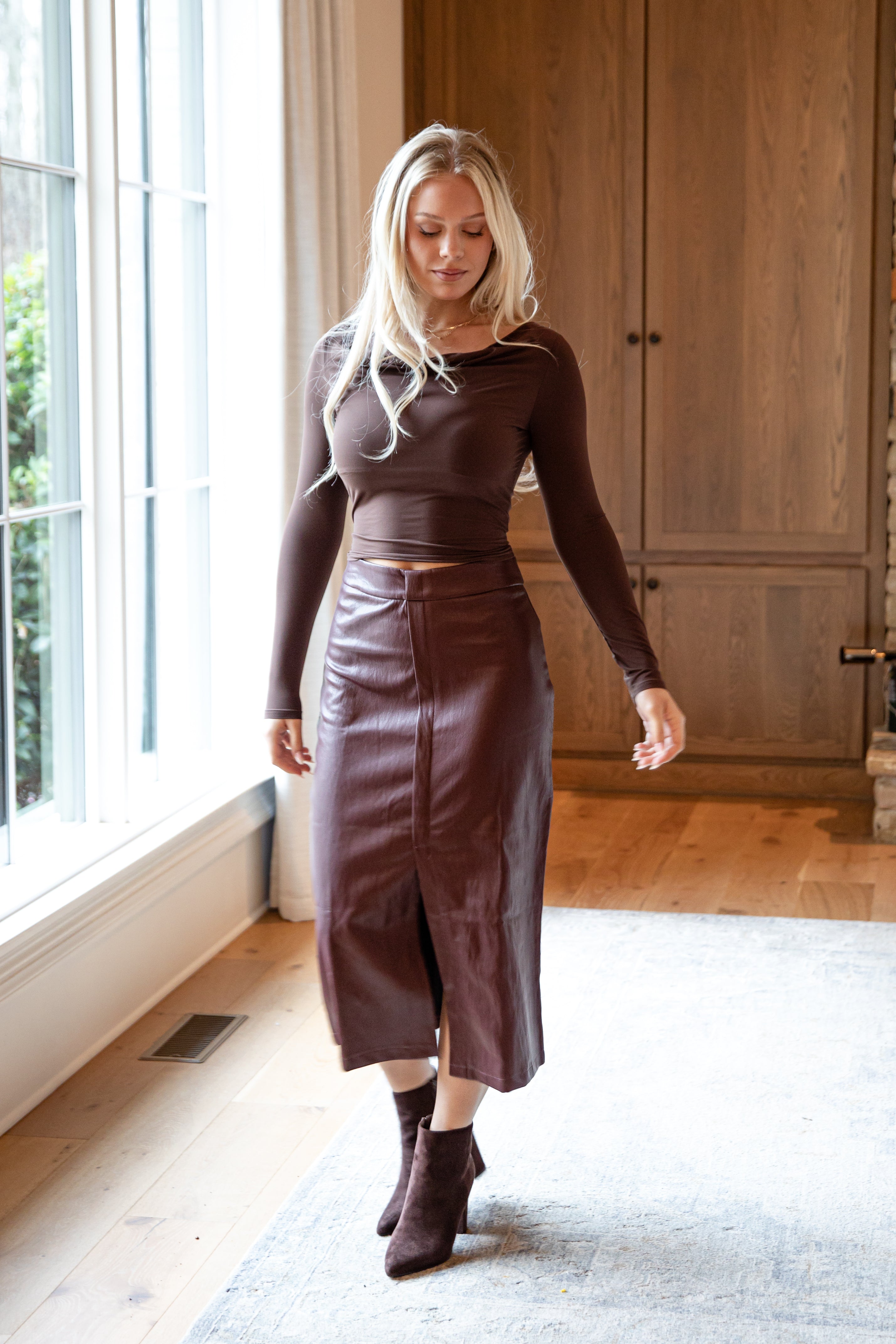 Woman wearing a brown top and leather skirt standing in a room with wooden flooring and large windows.