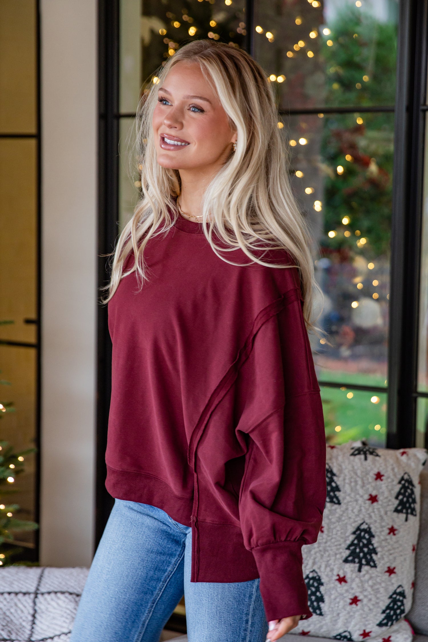 Woman wearing a maroon sweatshirt and blue jeans standing in front of a decorated window with lights.