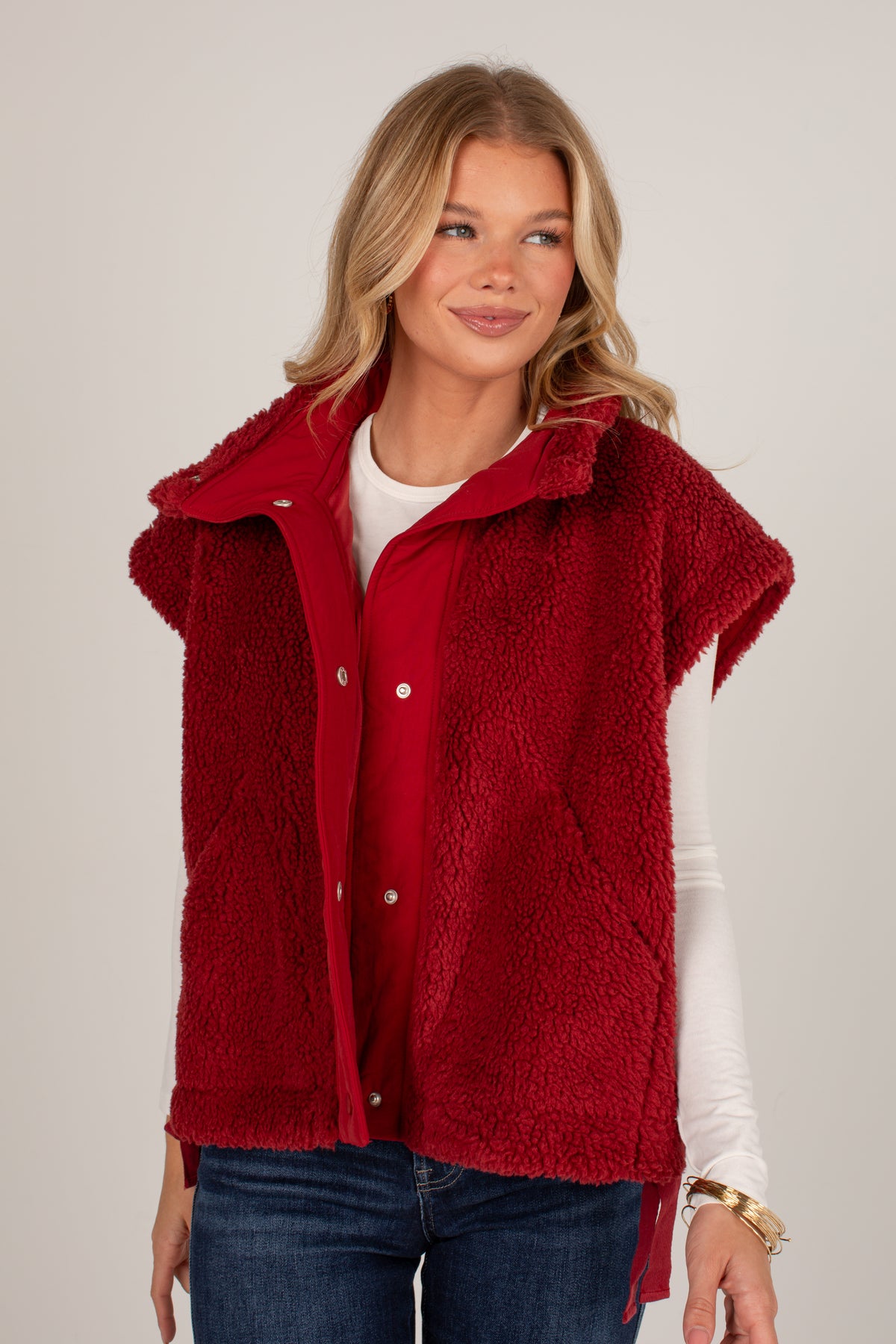 A woman wearing a sleeveless, plush red sherpa fleece vest over a white long-sleeve shirt and blue jeans, photographed against a neutral light gray studio background.