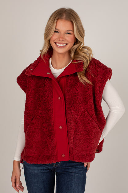 A smiling blonde woman wearing a red sherpa fleece button-front vest over a white long-sleeve top and blue jeans, photographed against a plain light gray studio background.
