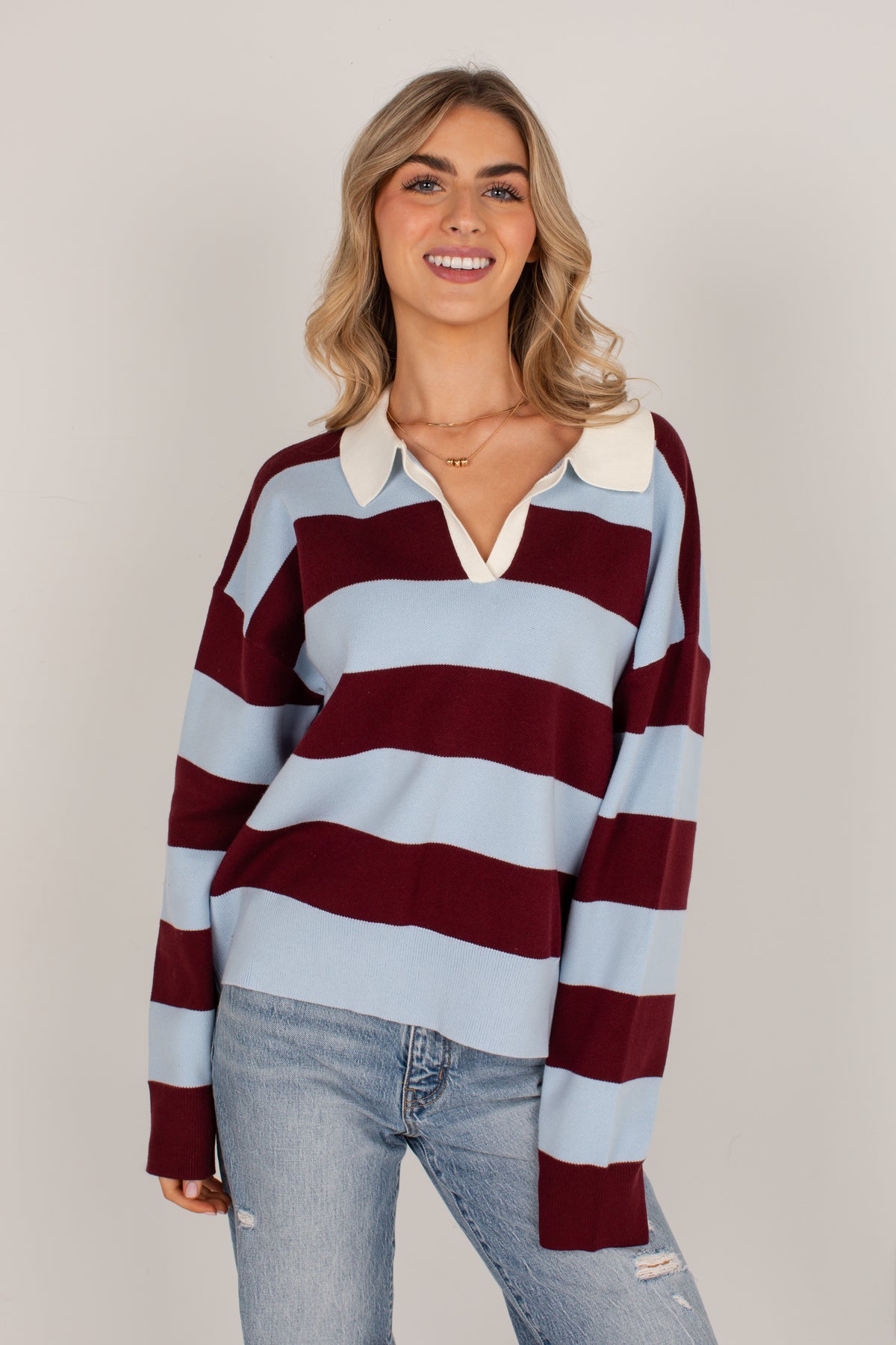 A woman modeling a long-sleeve rugby-style sweater with horizontal stripes in light blue, burgundy, and white, paired with light-wash jeans against a plain light gray studio background.