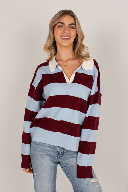 A woman modeling a long-sleeve rugby-style sweater with horizontal stripes in light blue, burgundy, and white, paired with light-wash jeans against a plain light gray studio background.