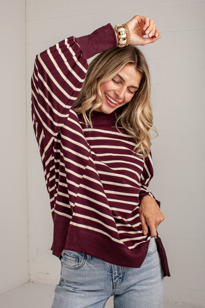 A woman wearing a burgundy and cream striped turtleneck sweater with wide sleeves and side ties, paired with light blue jeans, posing against a white brick wall.