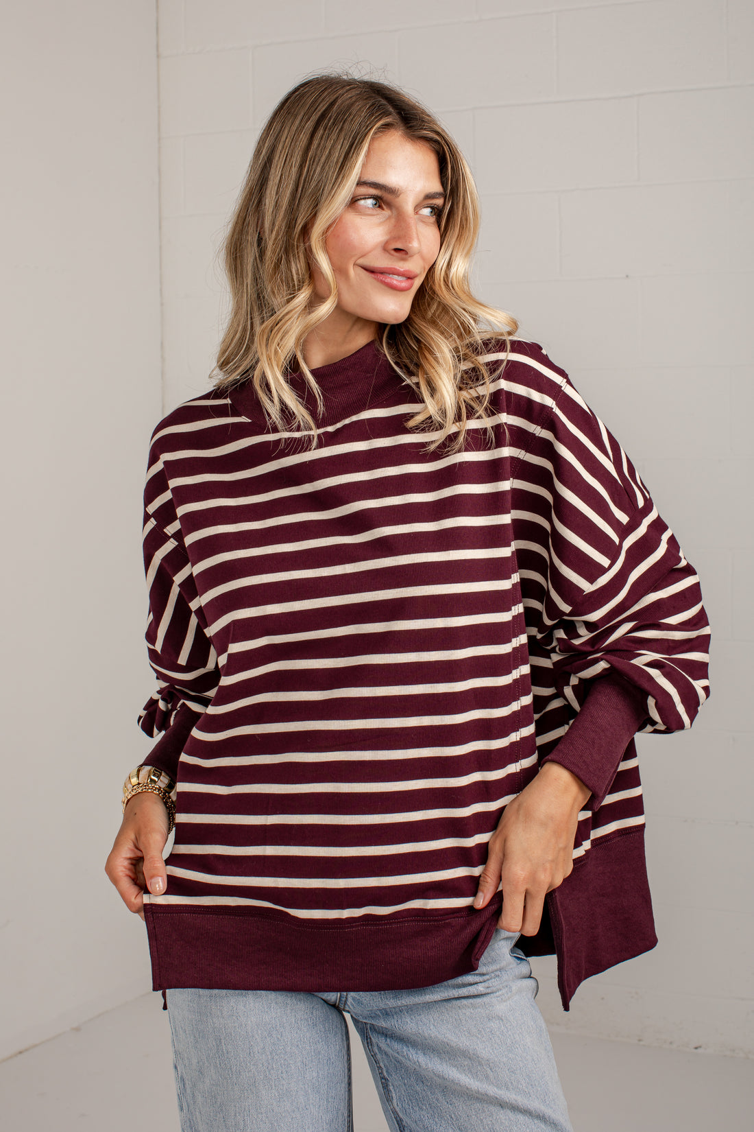 A woman wearing a loose-fitting, long-sleeve striped sweater in burgundy and cream, paired with light-wash jeans, standing against a white brick wall.