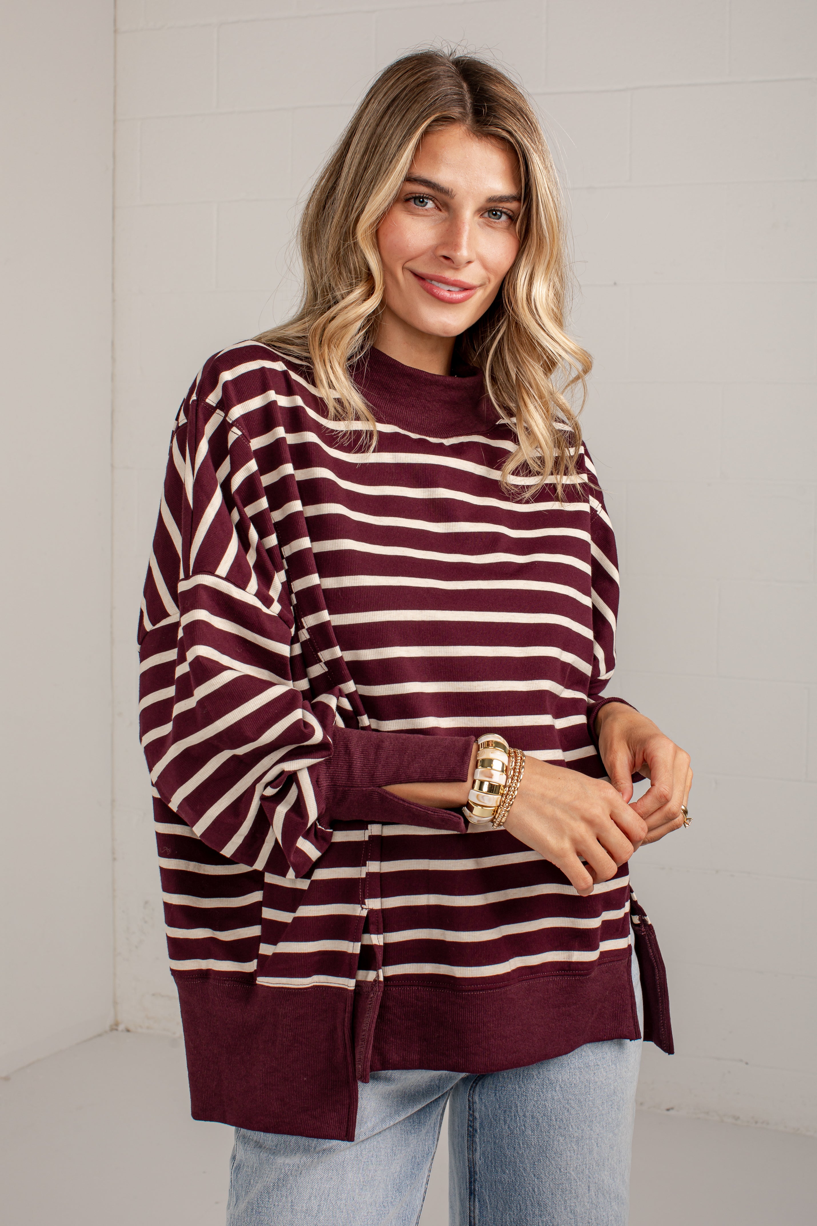 A woman wearing a burgundy and cream striped long-sleeve knit top with side slits, paired with light blue jeans, standing against a white brick wall.