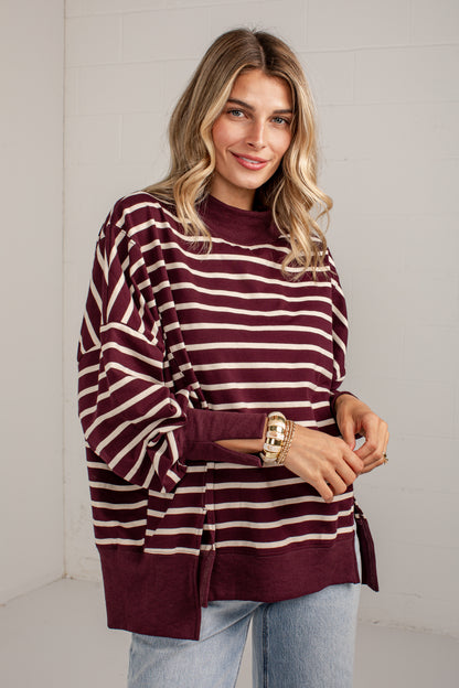 A woman wearing a burgundy and cream striped long-sleeve knit top with side slits, paired with light blue jeans, standing against a white brick wall.