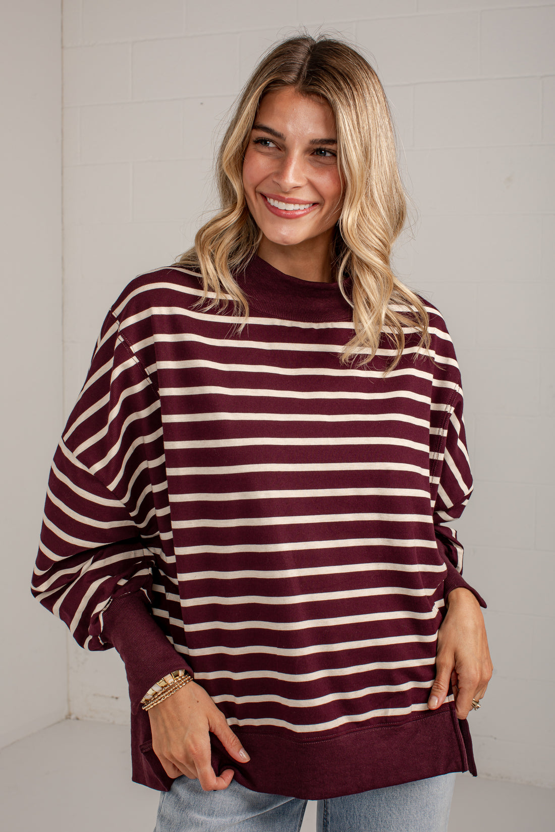 A woman wearing a burgundy and cream striped long-sleeve sweatshirt with a crew neck, paired with light blue jeans, standing against a white brick wall.