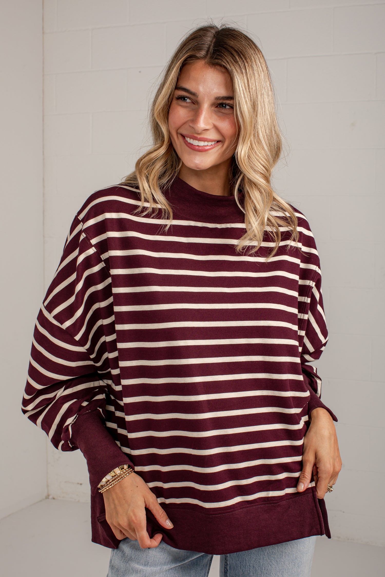 A woman wearing a burgundy and cream striped long-sleeve sweatshirt with a crew neck, paired with light blue jeans, standing against a white brick wall.