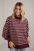 A woman wearing a burgundy and cream striped long-sleeve sweatshirt with a crew neck, paired with light blue jeans, standing against a white brick wall.