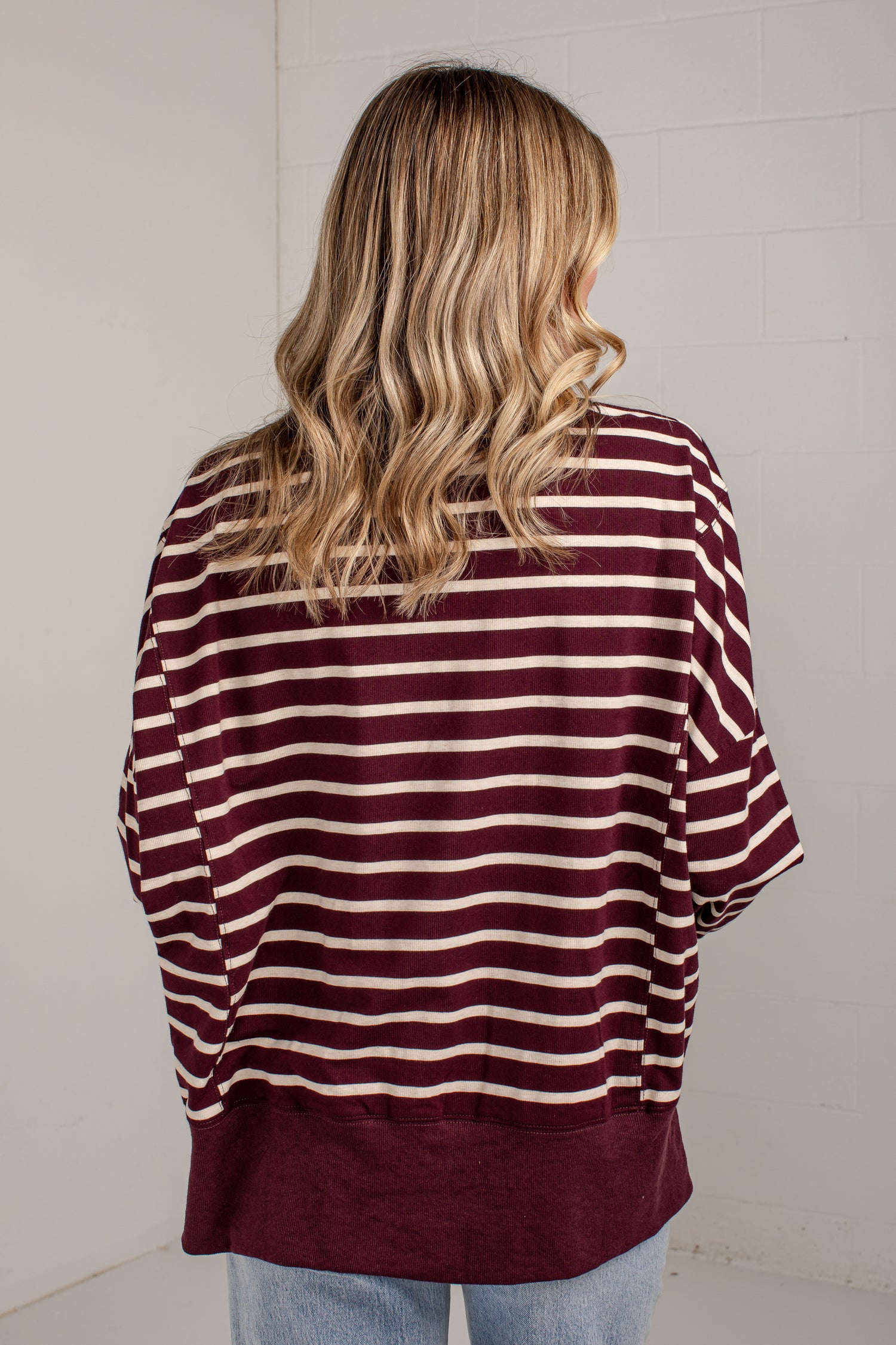 A woman viewed from behind wearing a loose-fitting short-sleeve top with horizontal burgundy and cream stripes, paired with light blue jeans, against a white brick wall.