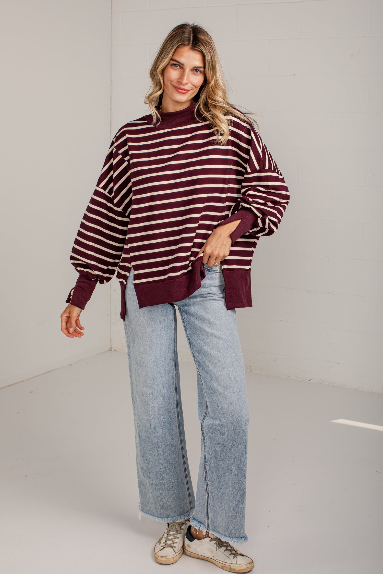 A woman modeling a maroon and white horizontally striped knit sweater with balloon sleeves, paired with light-wash wide-leg jeans and beige sneakers, in a bright white studio setting.