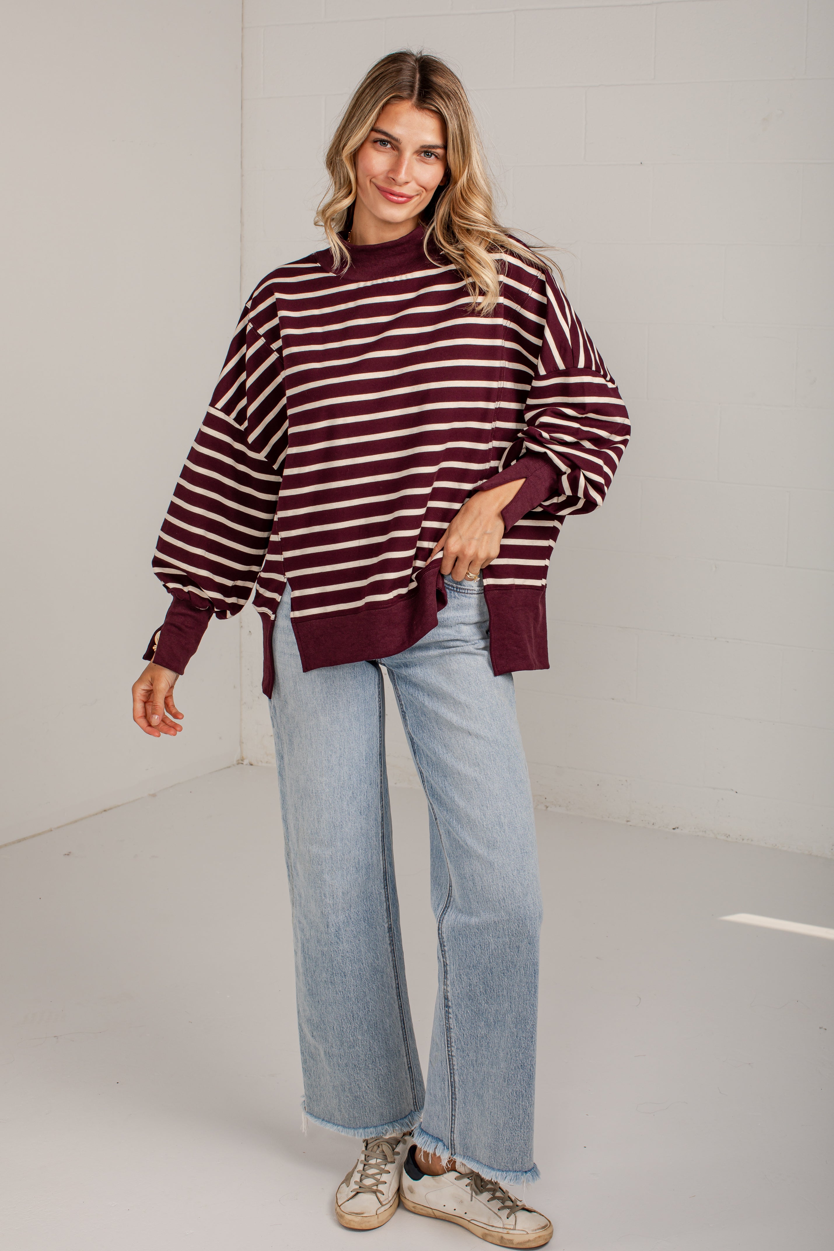 A woman modeling a maroon and white horizontally striped knit sweater with balloon sleeves, paired with light-wash wide-leg jeans and beige sneakers, in a bright white studio setting.
