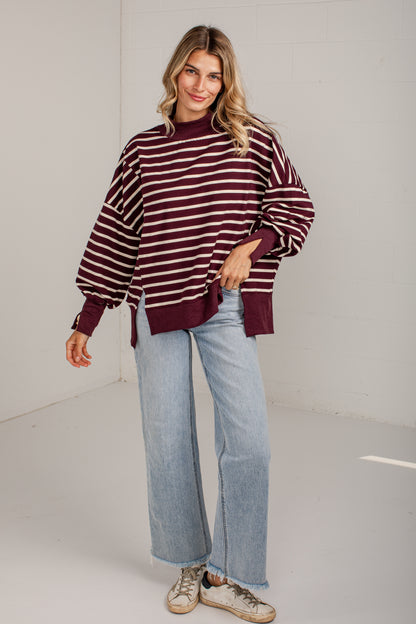 A woman modeling a maroon and white horizontally striped knit sweater with balloon sleeves, paired with light-wash wide-leg jeans and beige sneakers, in a bright white studio setting.