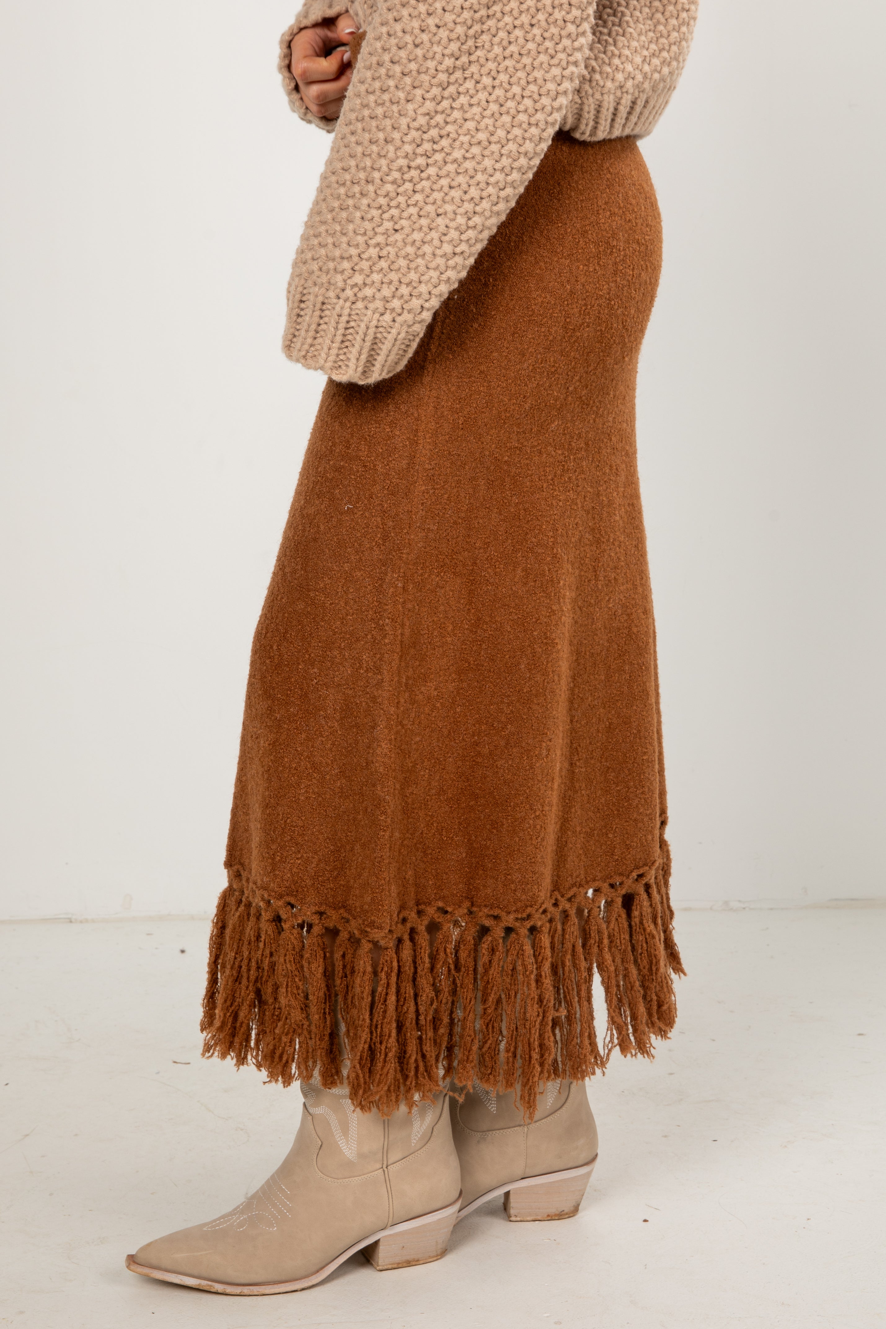 Brown fringed skirt worn with a beige sweater and boots on a white background