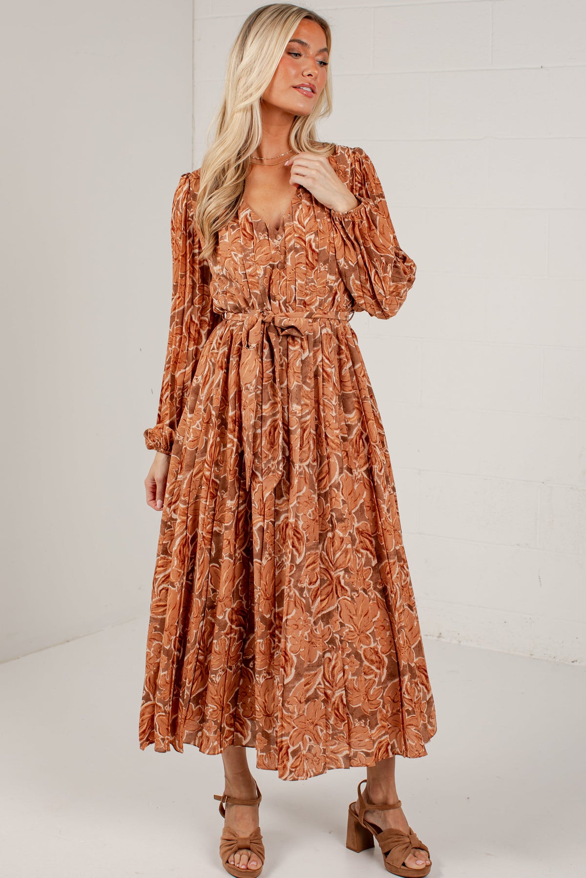 A woman modeling a rust-orange long-sleeve midi dress with a brown abstract floral pattern, featuring a tie waist and puffed sleeves, set against a white brick wall background.