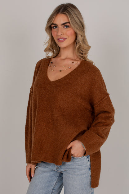 A woman wearing a fuzzy, oversized brown V-neck sweater and light blue jeans, standing against a solid light gray studio background.