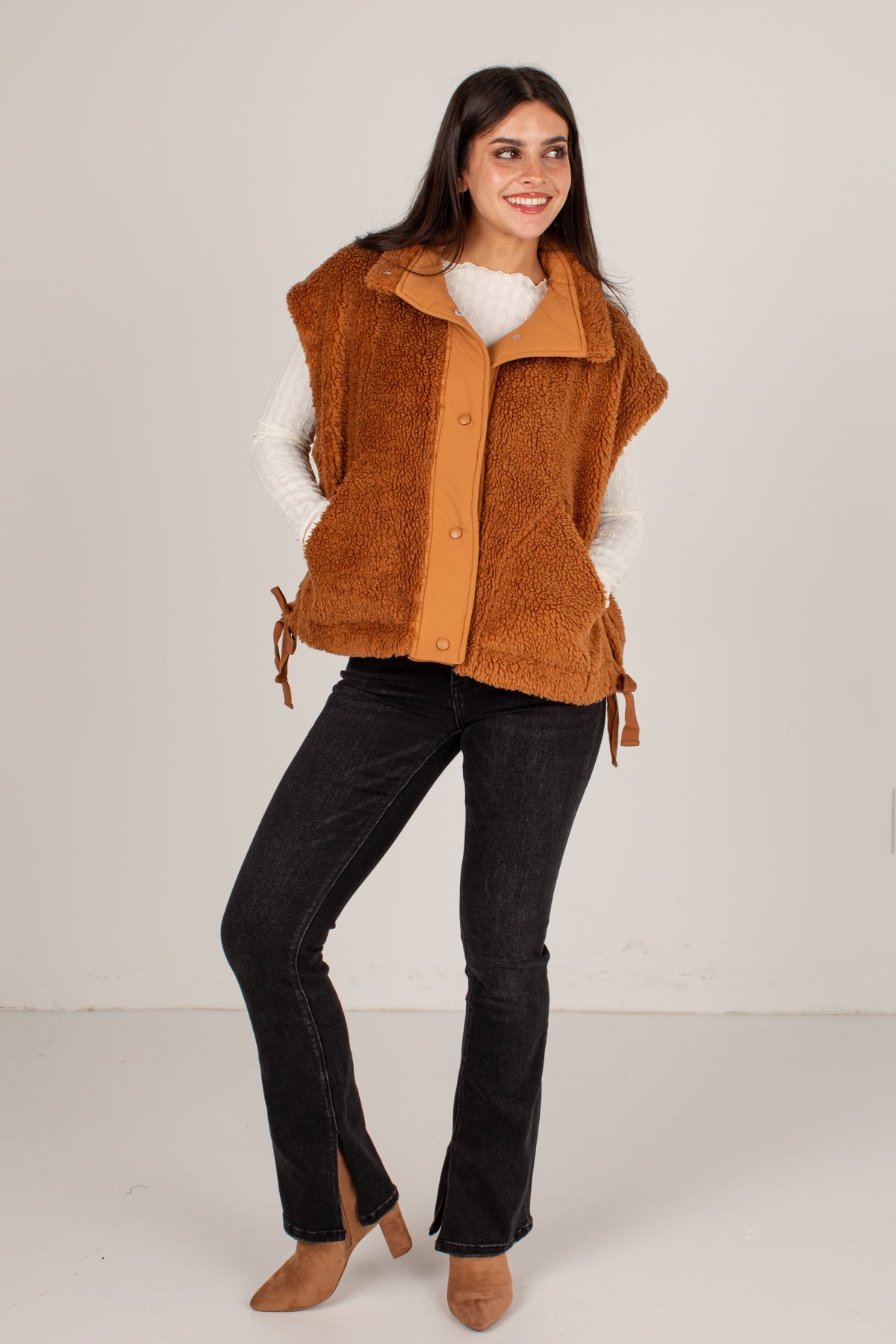 A woman modeling a brown sherpa fleece vest with a tan inner lining, worn over a white long-sleeve top and black flared jeans, against a plain light gray studio background.