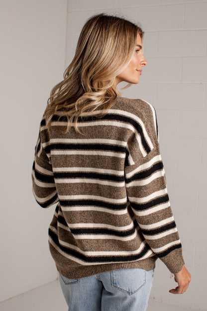 A woman wearing a brown, black, and cream striped knit sweater, shown from a three-quarters back view against a light gray brick wall.