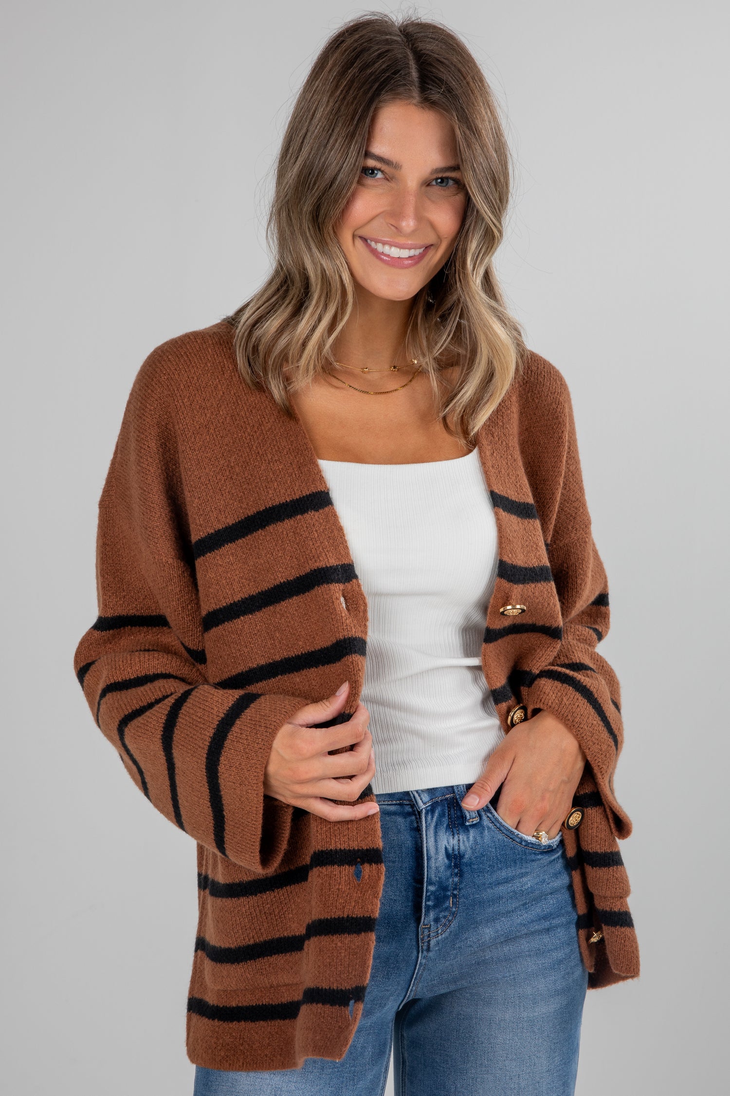 Woman wearing a brown cardigan with black stripes, white top, and blue jeans on a gray background