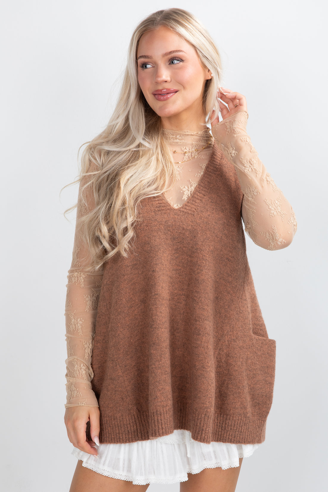 Woman wearing a brown knitted vest over a sheer top with a white skirt on a light gray background