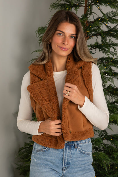 Woman wearing a brown vest and blue jeans standing in front of a Christmas tree.