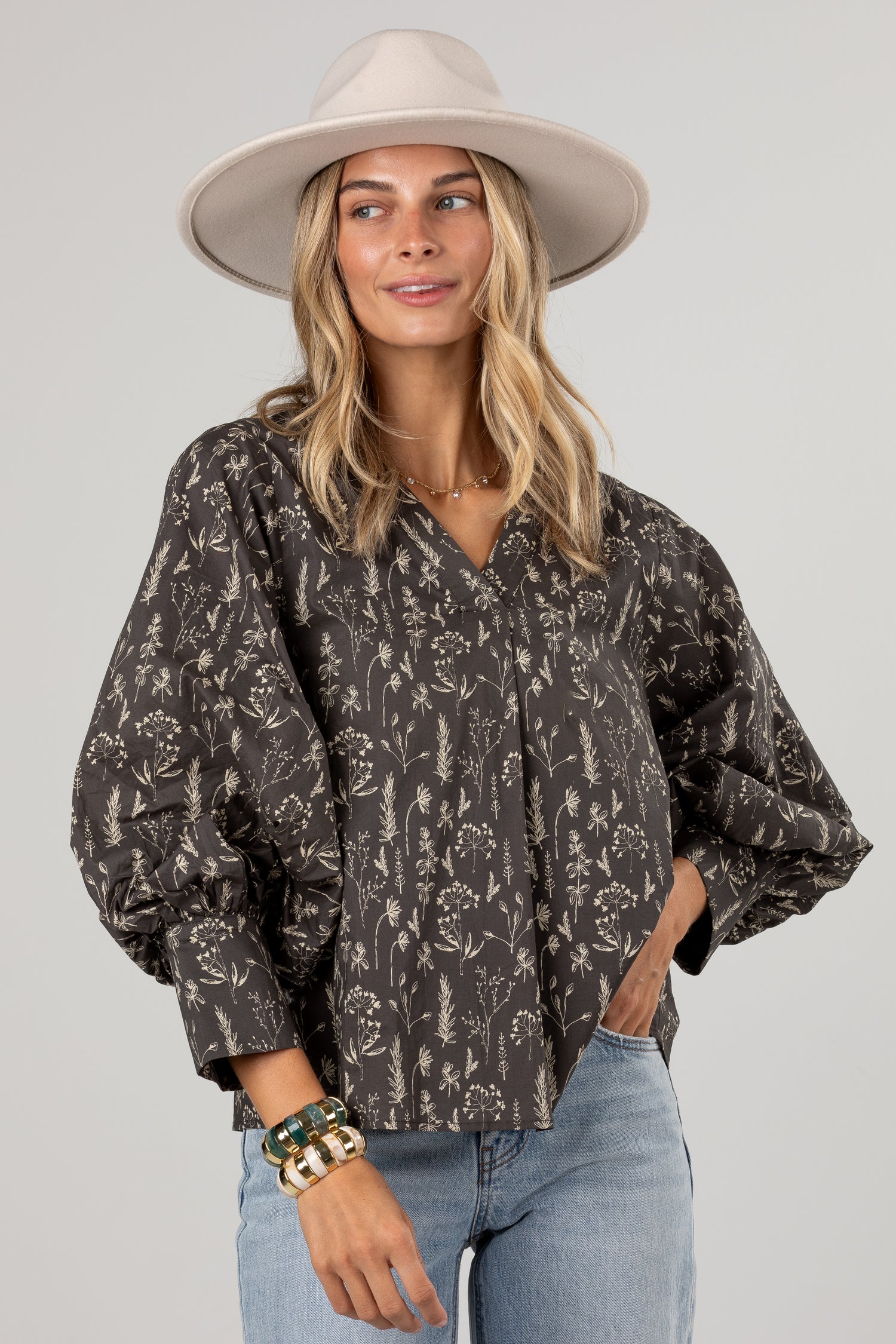 A woman wearing a dark brown and beige floral print blouse with voluminous balloon sleeves, paired with light blue jeans and a beige wide-brim hat, against a solid light gray background.