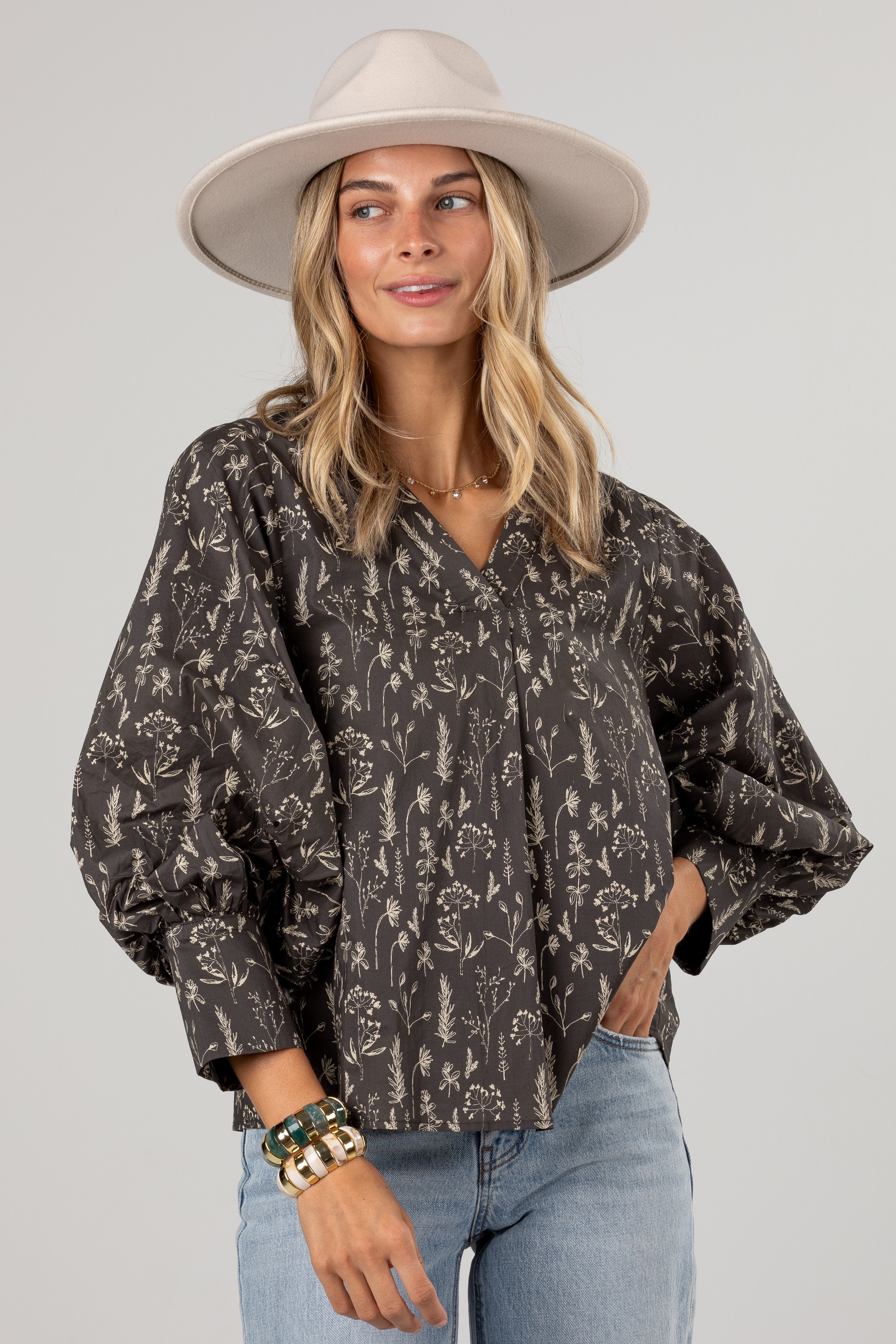 A woman wearing a dark brown and beige floral print blouse with voluminous balloon sleeves, paired with light blue jeans and a beige wide-brim hat, against a solid light gray background.