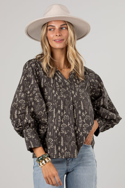 A woman wearing a dark brown and beige floral print blouse with voluminous balloon sleeves, paired with light blue jeans and a beige wide-brim hat, against a solid light gray background.