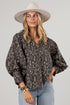 A woman wearing a dark brown and beige floral print blouse with voluminous balloon sleeves, paired with light blue jeans and a beige wide-brim hat, against a solid light gray background.