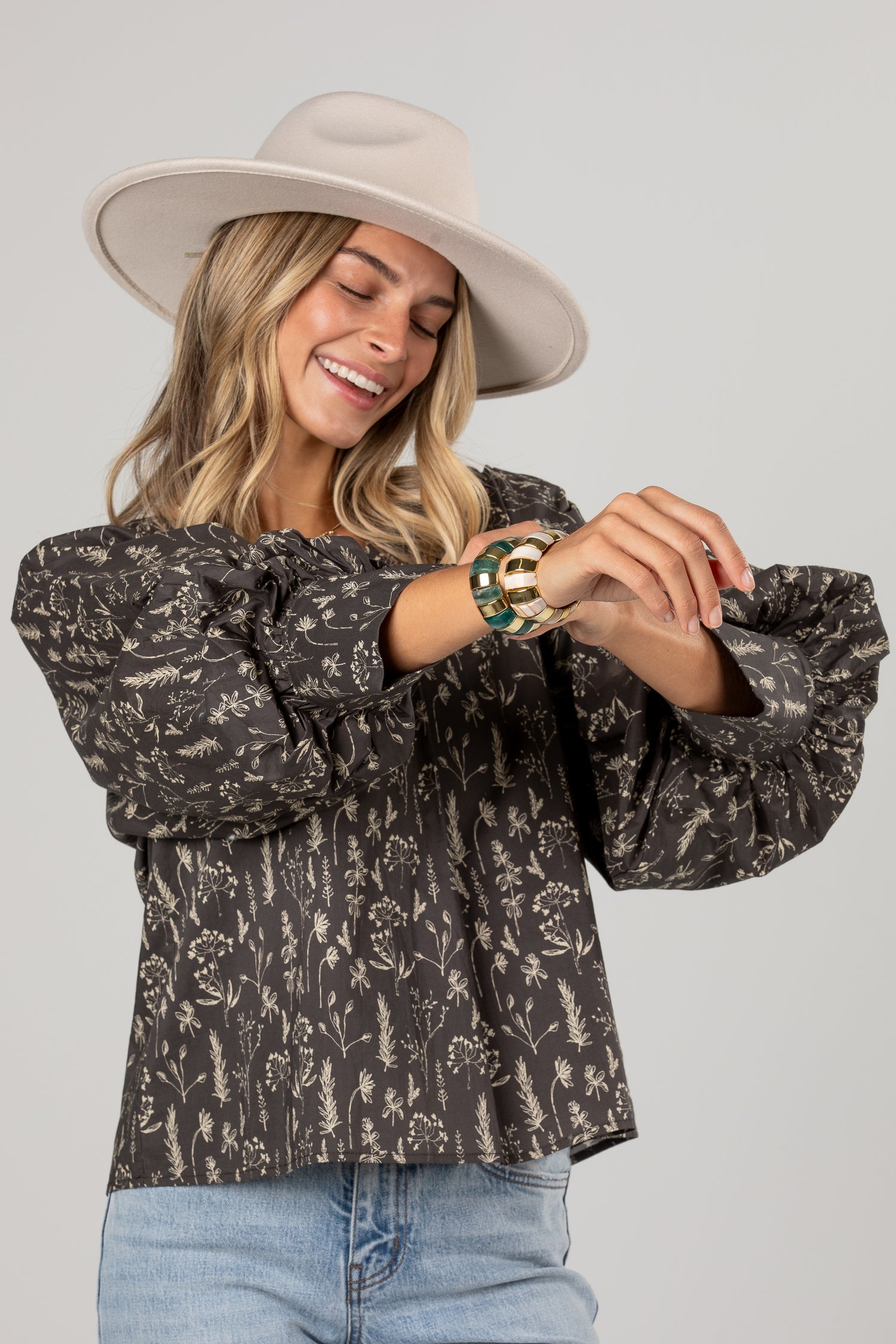 A woman wearing a dark brown and beige floral print blouse with dramatic puff sleeves, a wide-brimmed beige hat, and light blue jeans, smiling as she looks down at her wrist.