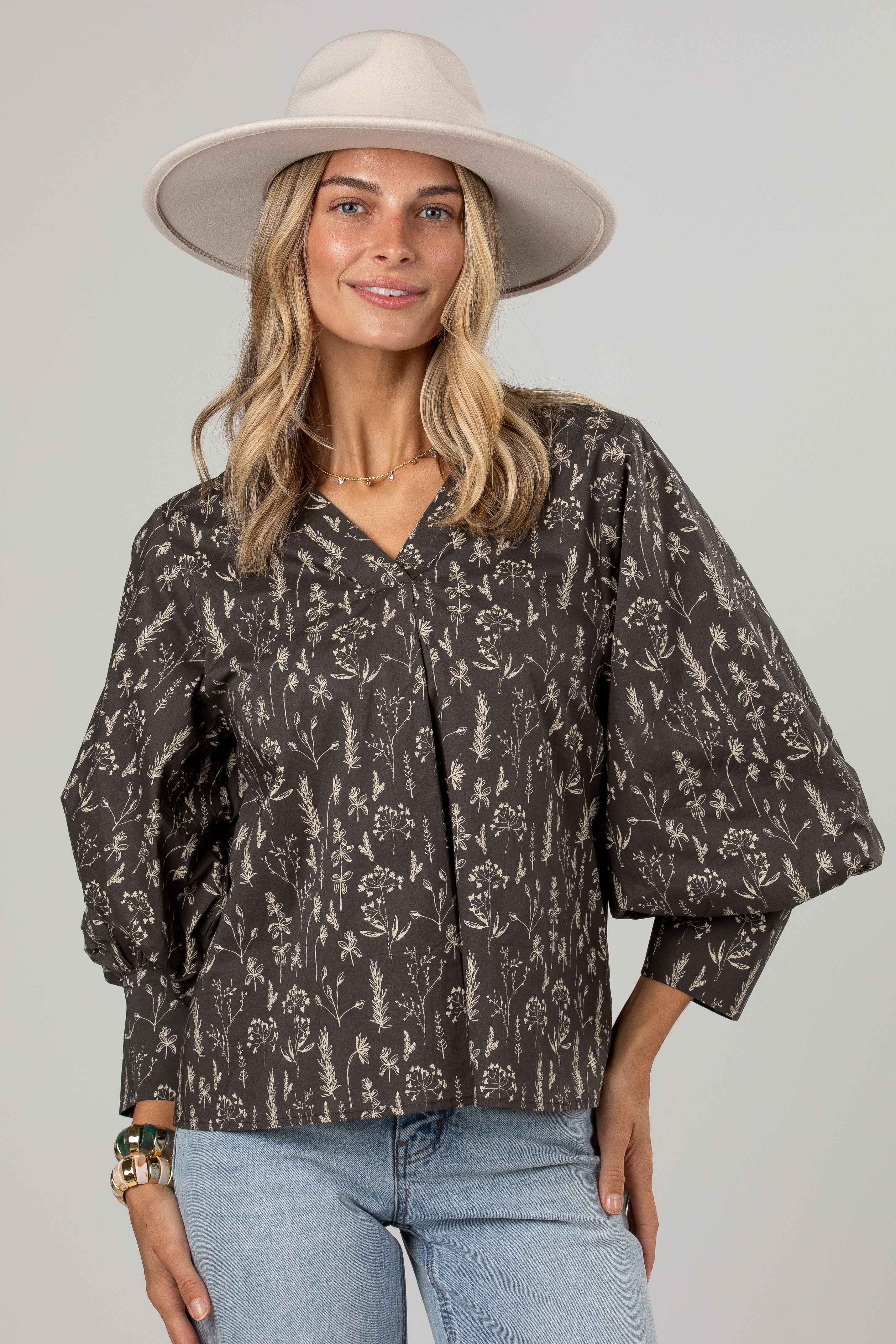 A woman wearing a dark brown and beige floral print blouse with voluminous sleeves, a wide-brimmed beige hat, and light blue jeans, photographed against a solid light gray background.