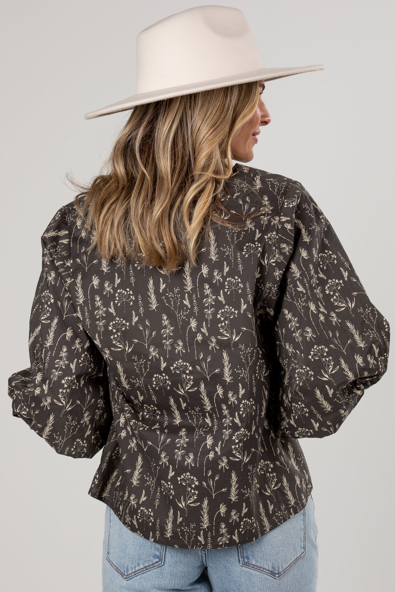 A woman shown from the back wearing a dark brown long-sleeve blouse with a beige botanical pattern, paired with light blue jeans and a cream wide-brim hat, against a light gray studio background.