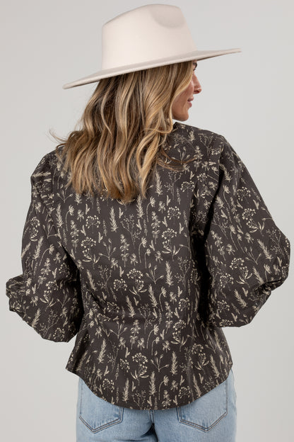 A woman shown from the back wearing a dark brown long-sleeve blouse with a beige botanical pattern, paired with light blue jeans and a cream wide-brim hat, against a light gray studio background.