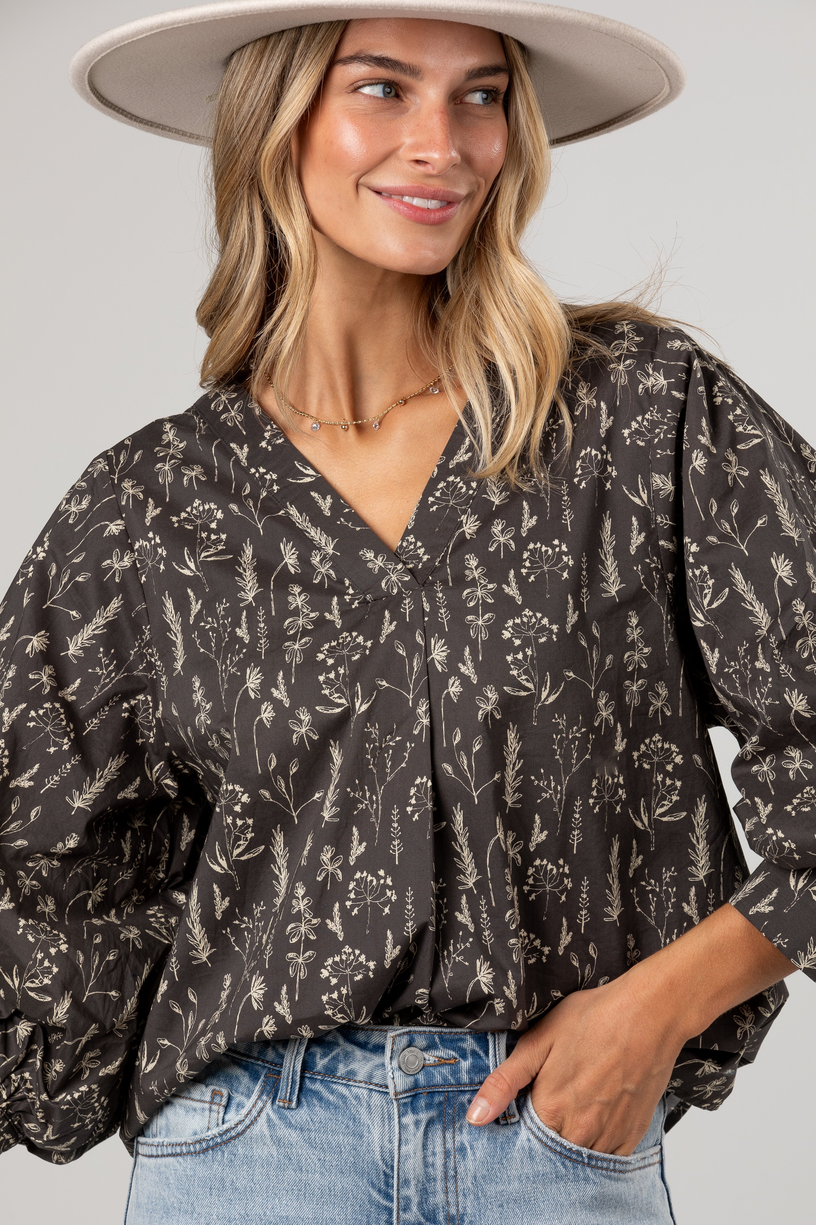 A woman wearing a dark brown floral print blouse with puff sleeves and a V-neck, paired with light blue jeans and a wide-brimmed beige hat, against a solid light gray background.