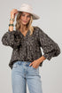 A woman modeling a dark gray and black floral print blouse with voluminous long sleeves, paired with light blue jeans and a cream wide-brim hat, against a solid light gray background.
