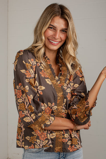 A woman wearing a brown floral blouse with orange and beige botanical patterns, paired with light blue jeans, standing against a white paneled wall.