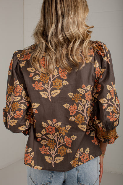 A woman viewed from the back wearing a brown long-sleeve blouse with a floral pattern in orange, beige, and muted pink, paired with light blue jeans.