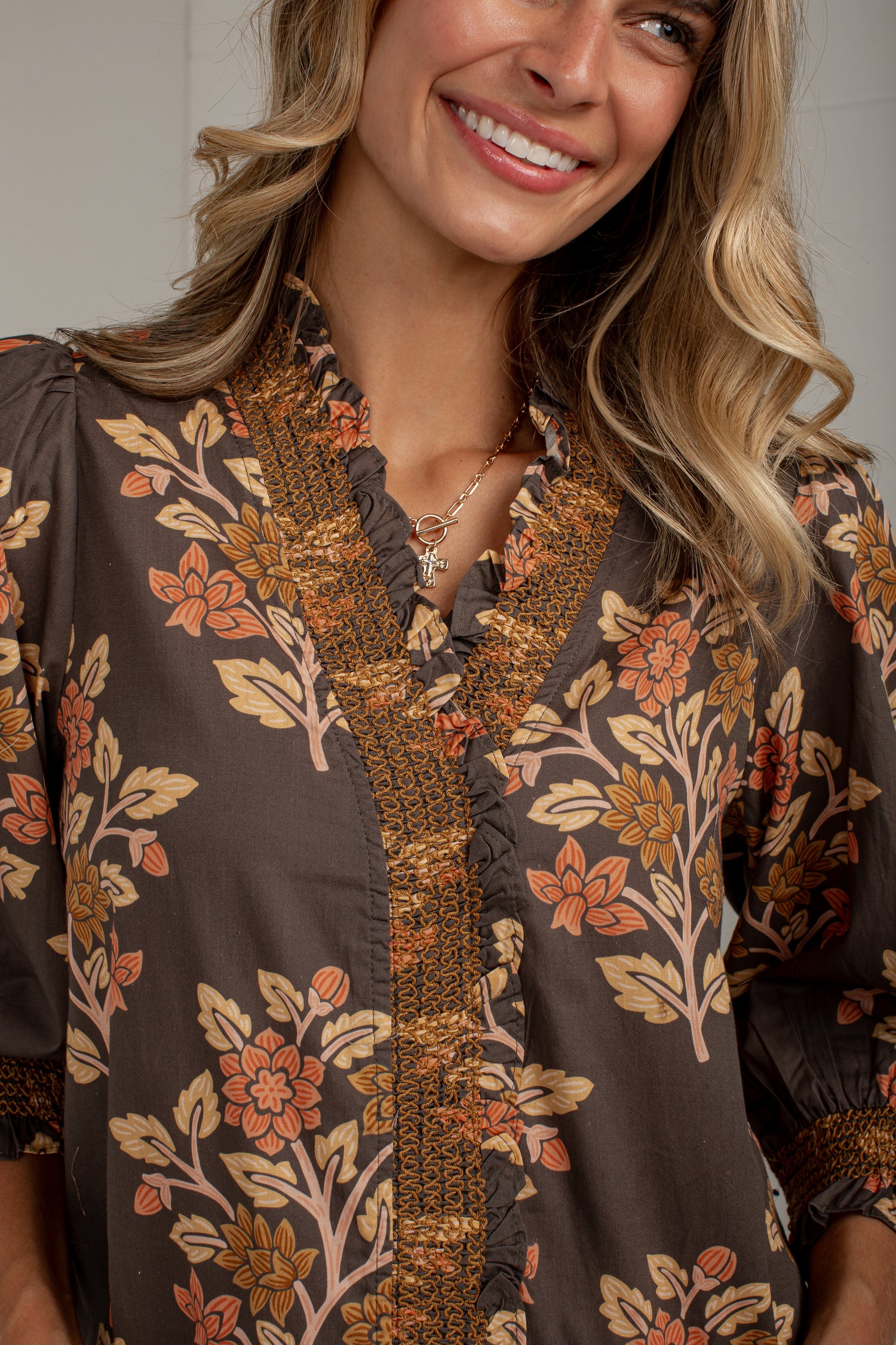 A close-up of a woman wearing a brown blouse with a floral print in orange, beige, and brown, featuring a ruffled V-neckline and embroidered trim.