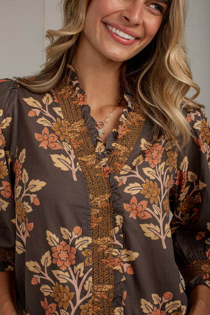 A close-up of a woman wearing a brown blouse with a floral print in orange, beige, and brown, featuring a ruffled V-neckline and embroidered trim.