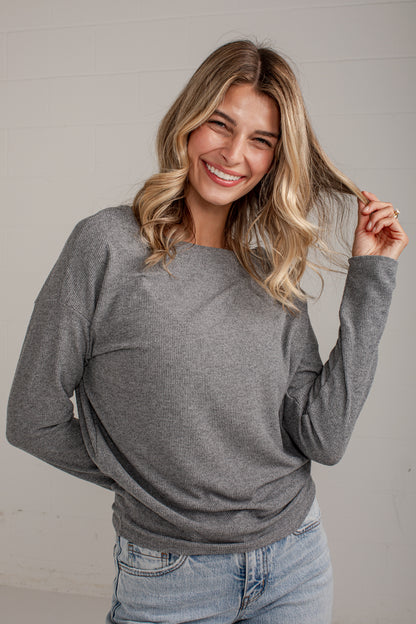 A smiling blonde woman wearing a heather gray long-sleeve crewneck sweater and light blue jeans, posing against a light gray paneled wall.