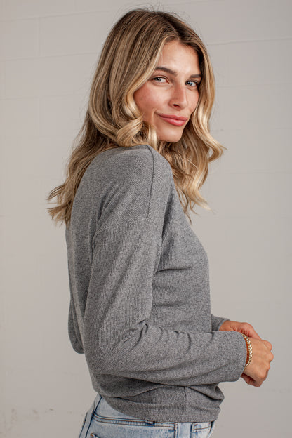 A woman with blonde wavy hair wearing a heather gray long-sleeve knit top and light blue jeans, photographed against a light gray wall.
