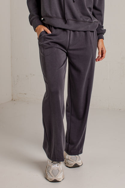 A person wearing dark charcoal gray sweatpants with a matching sweatshirt and white sneakers, standing in a minimalist studio setting with a white brick wall background.