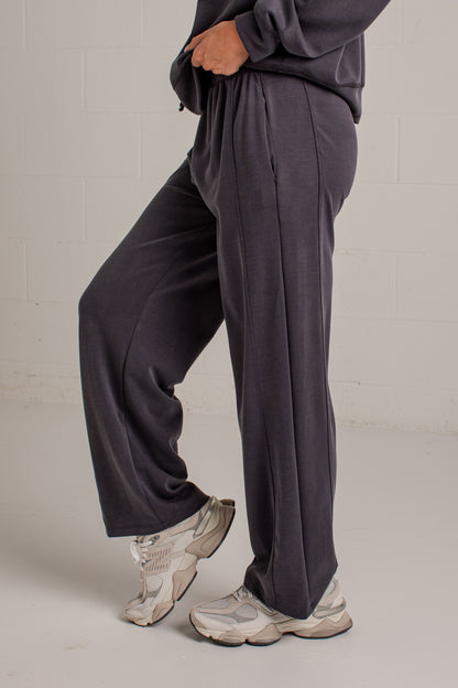 A person wearing loose, wide-leg dark charcoal sweatpants and chunky off-white sneakers, standing against a light gray brick wall.