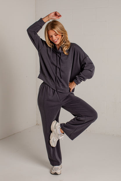 A woman modeling a dark gray two-piece loungewear set consisting of a cropped hoodie and matching sweatpants, paired with white sneakers, in a bright studio setting.