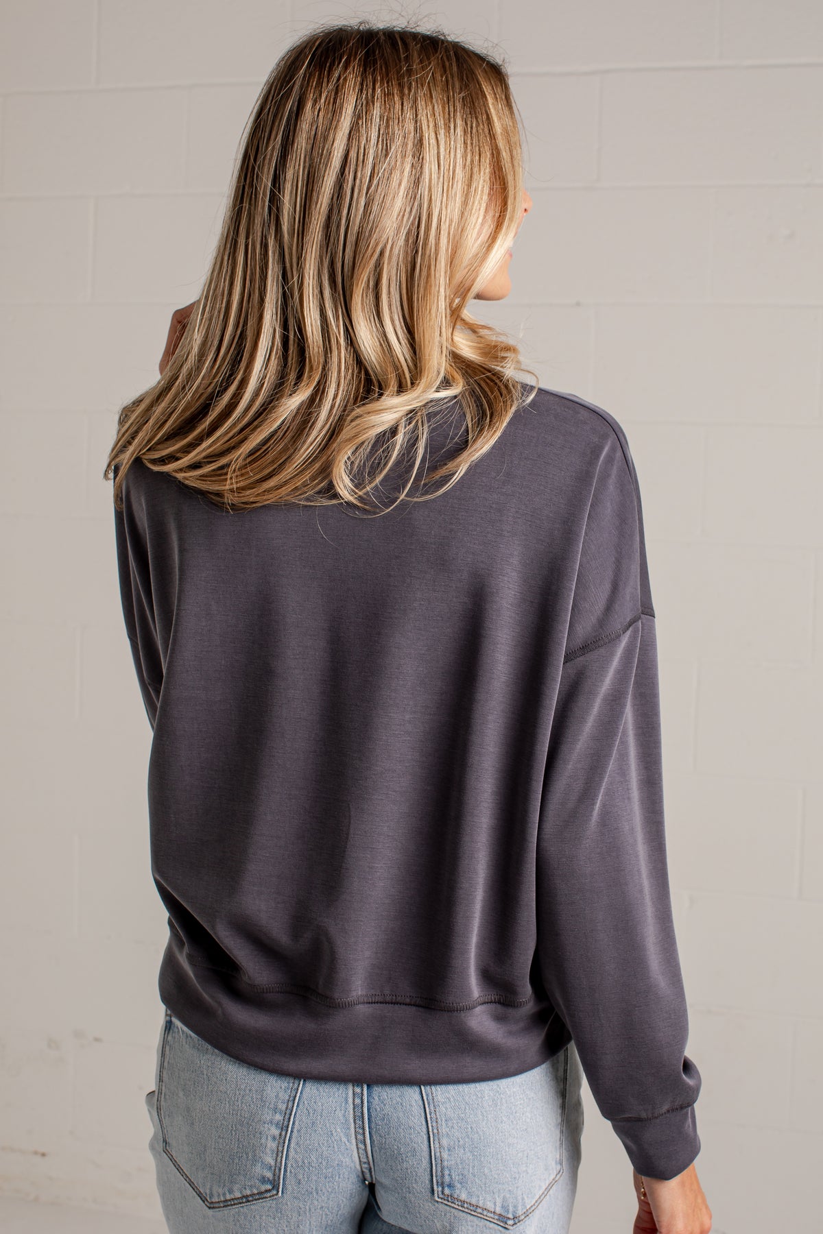 A woman viewed from the back wearing a dark charcoal gray crewneck sweatshirt and light blue jeans, standing against a light gray brick wall.