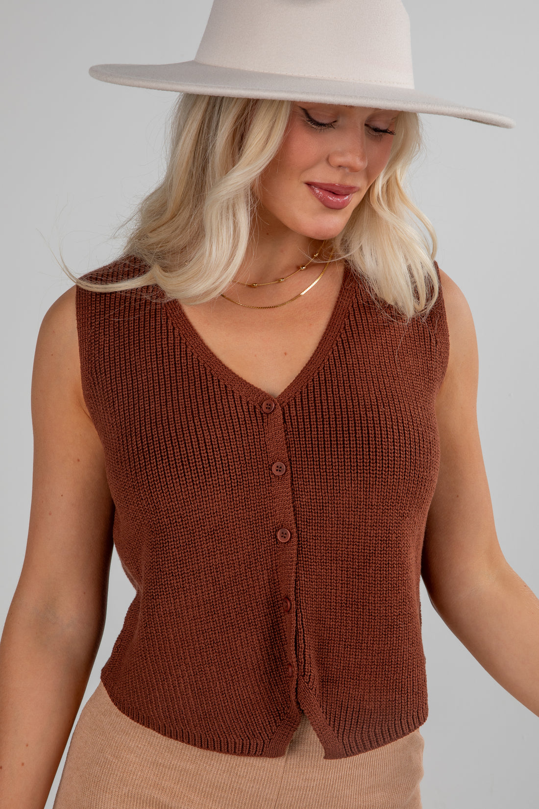 Woman wearing a brown knitted vest and beige hat against a plain background