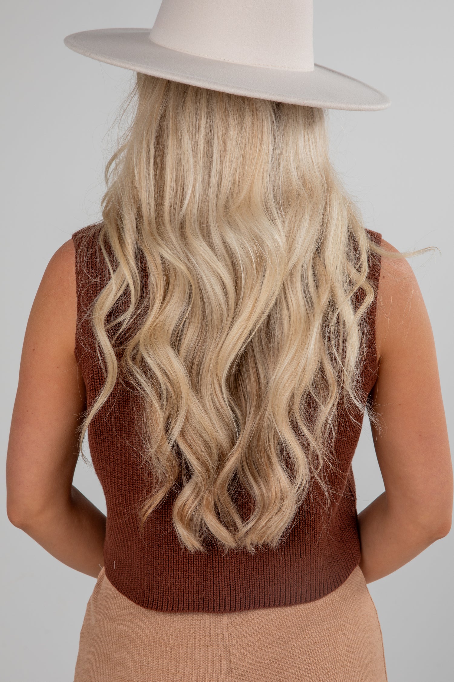 Person wearing a brown sleeveless top and beige hat with long blonde hair on a plain background