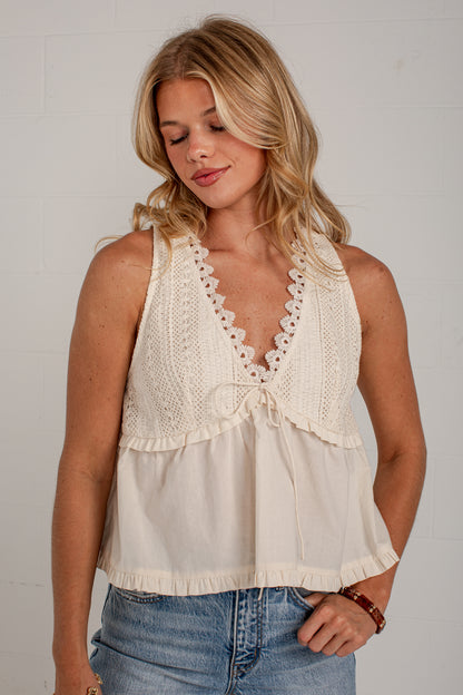 A woman wearing a cream-colored sleeveless crochet top with a ruffled V-neckline and tie detail, paired with blue jeans, standing against a white brick wall.