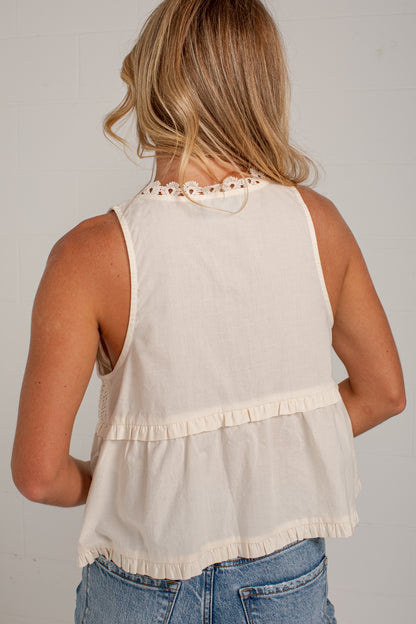 A woman viewed from the back wearing a sleeveless cream-colored ruffled top and blue jeans against a light gray wall.