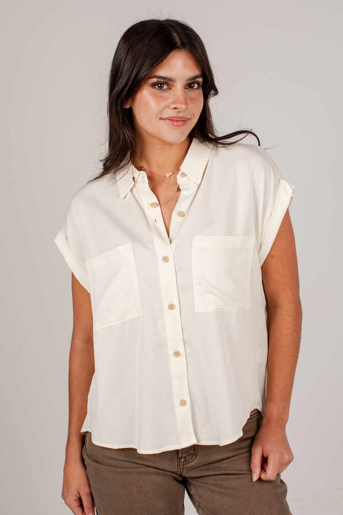 A woman wearing a short-sleeve button-up shirt in off-white with two chest pockets, paired with brown pants, against a plain light gray studio background.